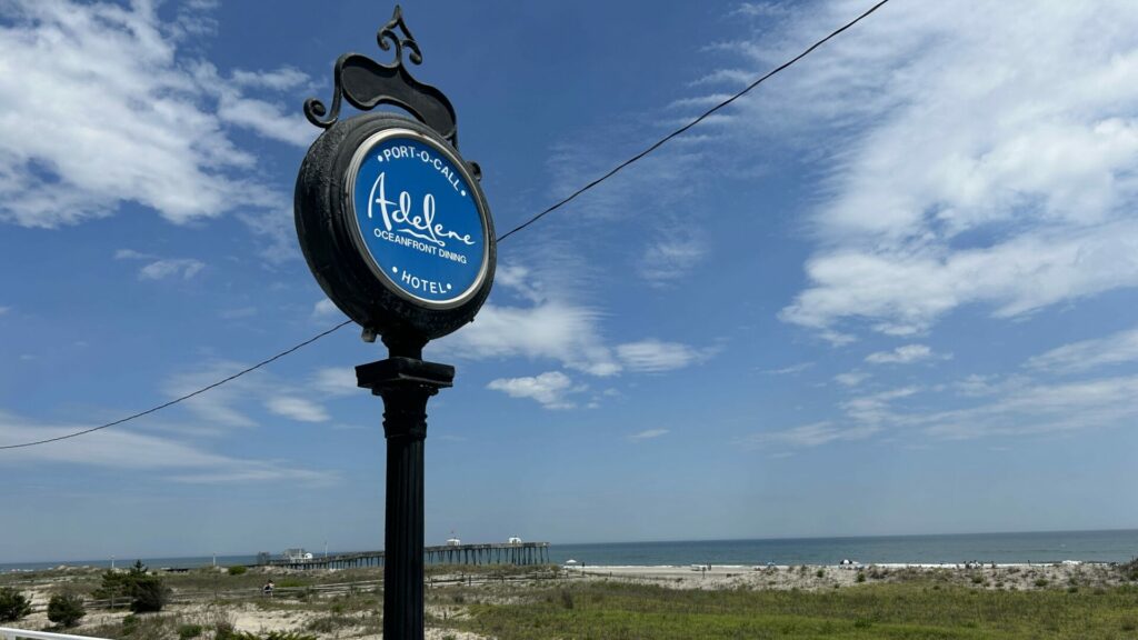 A black wrought iron sign that reads "Port-O-Call Adelene Oceanfront Dining" facing the boardwalk with the ocean, beach, and pier in the background