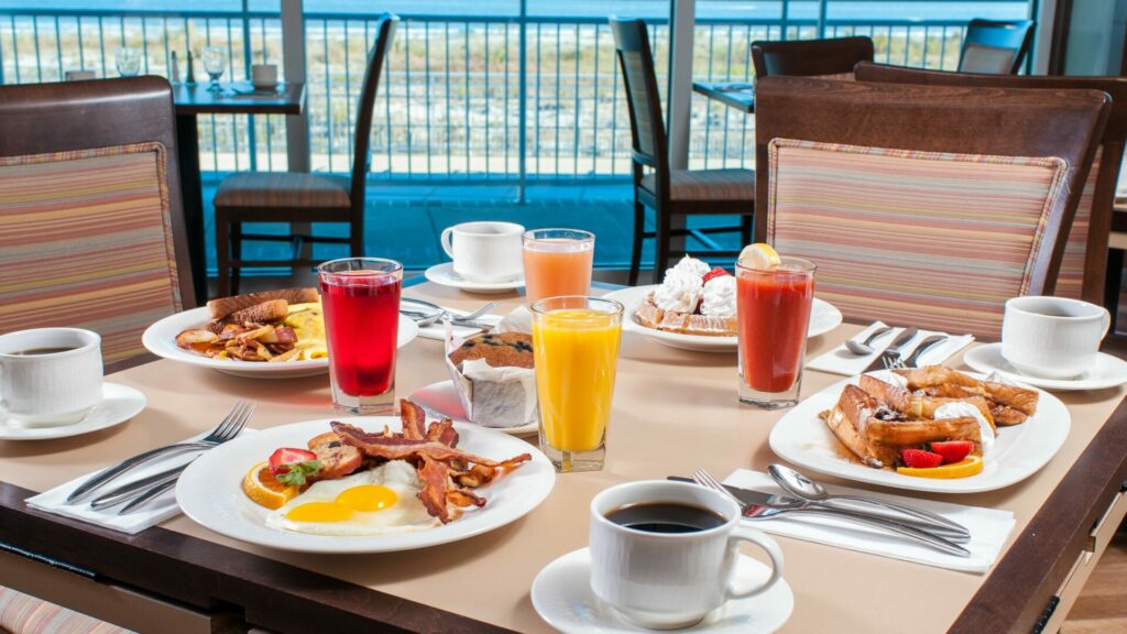 A table full of breakfast food, mocktails, and coffee. The beach and ocean can be seen through the windows behind it.
