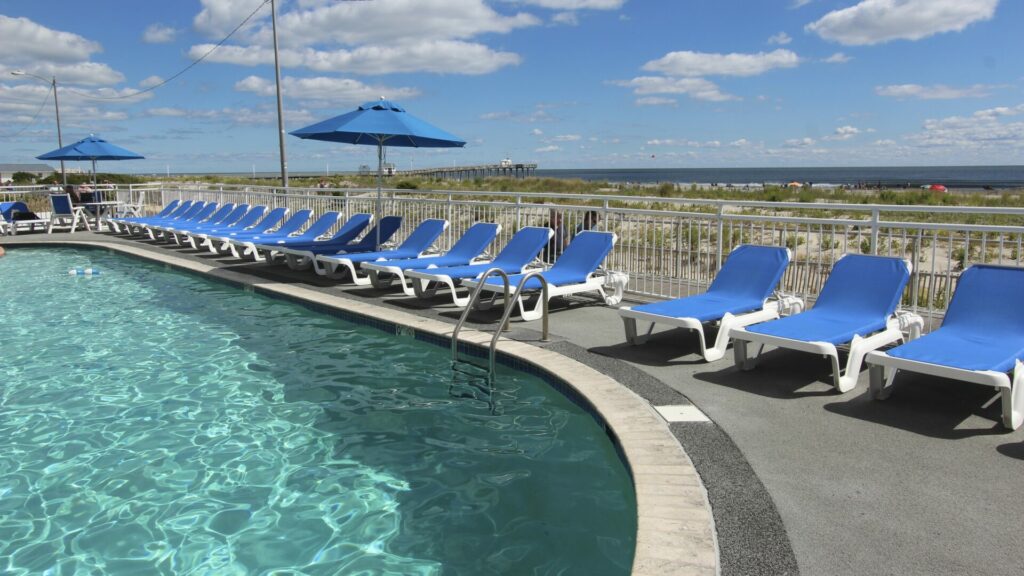 A pool with several blue lounge chairs surrounding it. Outside the fence, you can see the beach and ocean.