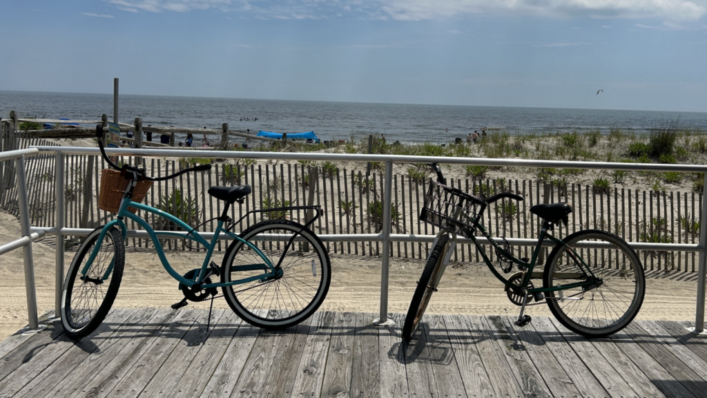 Two bikes on the boardwalk propped up against a railing in front of the ocean and beach.