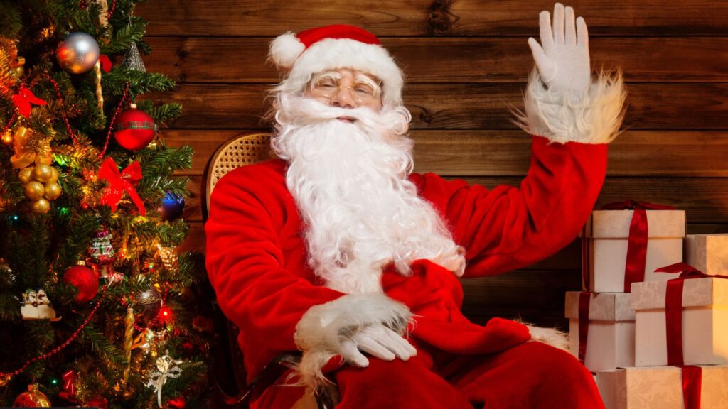 Santa Claus dressed in his red suit next to a Christmas tree smiling and waving